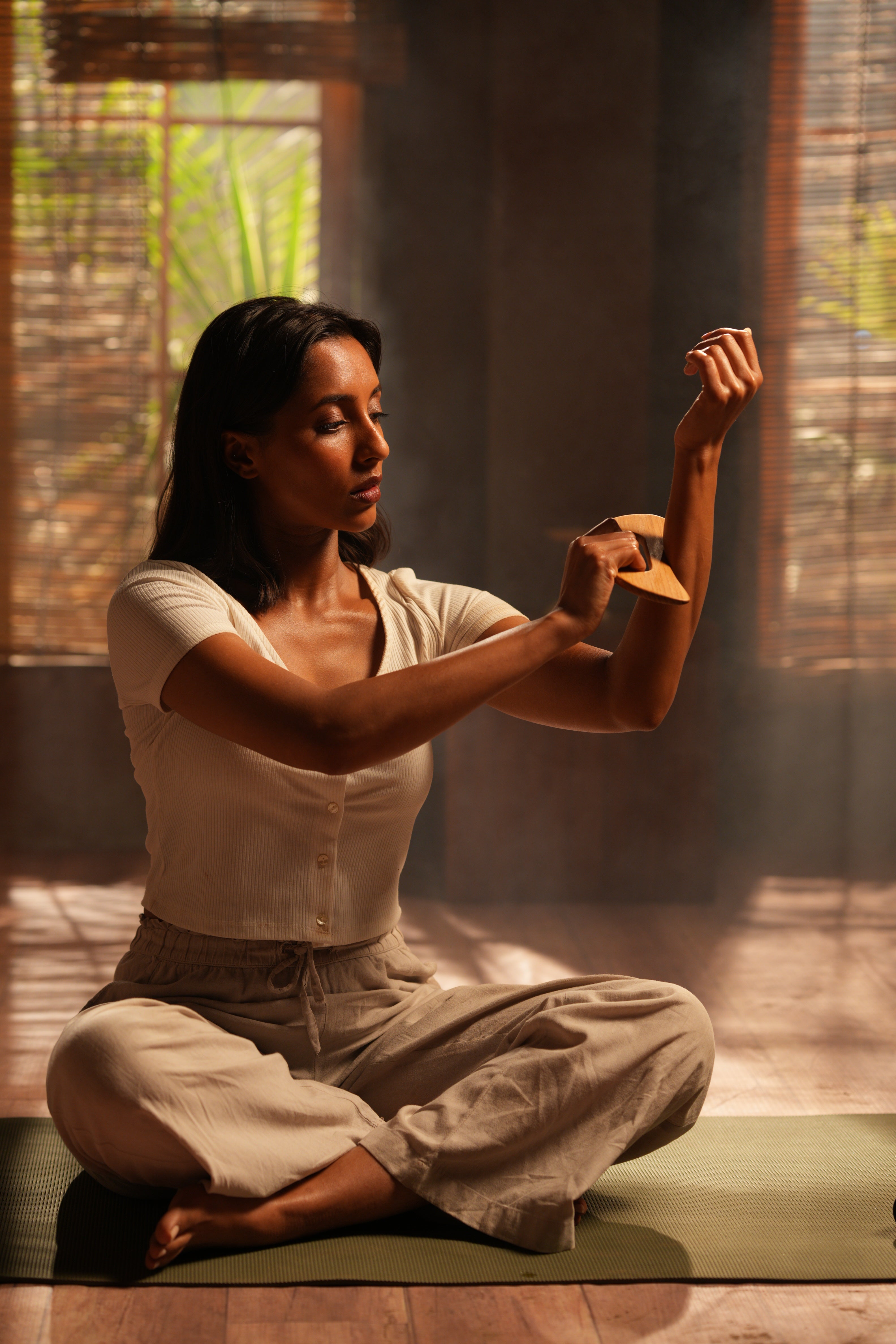 Woman in a white shirt and beige pants sitting and using sculpting gua sha tool with body oil