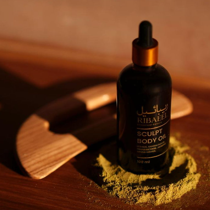 Black bottle of sculpt body oil with a wooden gua sha applicator on a wooden surface.